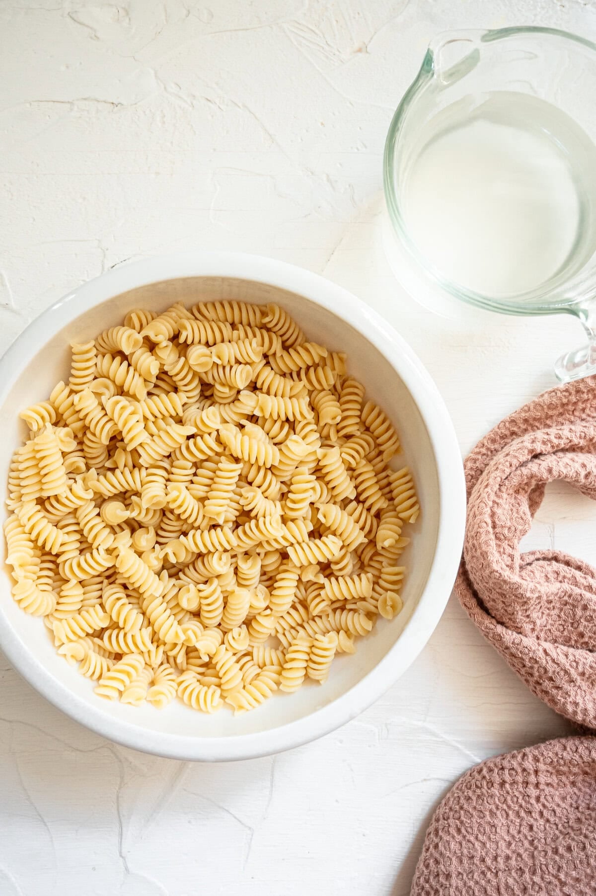 A bowl of cooked rotini noodles next to a glass pitcher with pasta water.