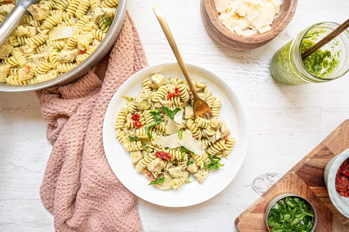 A skillet of creamy chicken pesto pasta next to a plate of it.
