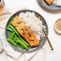 A baked Asian salmon filet on a plate with snap peas and rice.