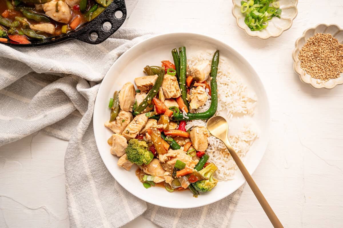 Stir-fried chicken and veggies on a plate with rice topped with sesame seeds and sliced green onions.