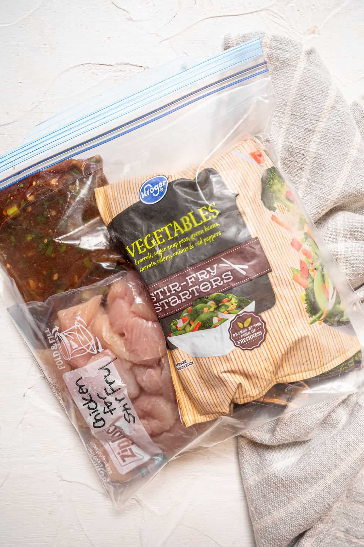 A gallon size freezer bag labeled chicken stir fry with a smaller freezer bag of raw chicken pieces, a freezer bag of homemade stir dry sauce, and a store-bought bag of frozen stir fry veggies.