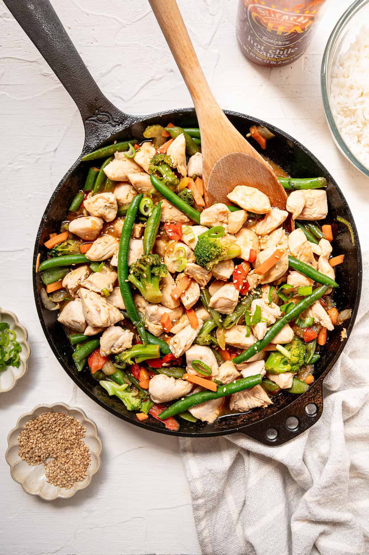 Stir fried chicken and veggies being served from a cast iron skillet with small bowls of sesame seeds and sliced green onions next to it.