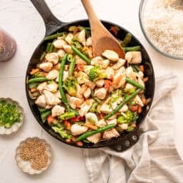 A cast iron skillet full of chicken stir fry with bowls of rice, sliced green onions, and sesame seeds around it.