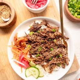 Korean beef on a plate with white rice, cucumber slices, shredded carrots, pickled red onions, and Kimchi on the side.
