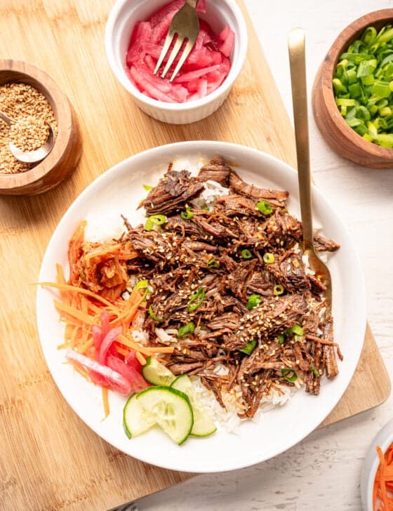 Korean beef on a plate with white rice, cucumber slices, shredded carrots, pickled red onions, and Kimchi on the side.