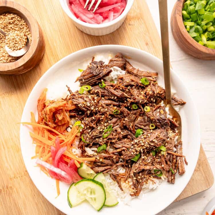 Korean beef on a plate with white rice, cucumber slices, shredded carrots, pickled red onions, and Kimchi on the side.