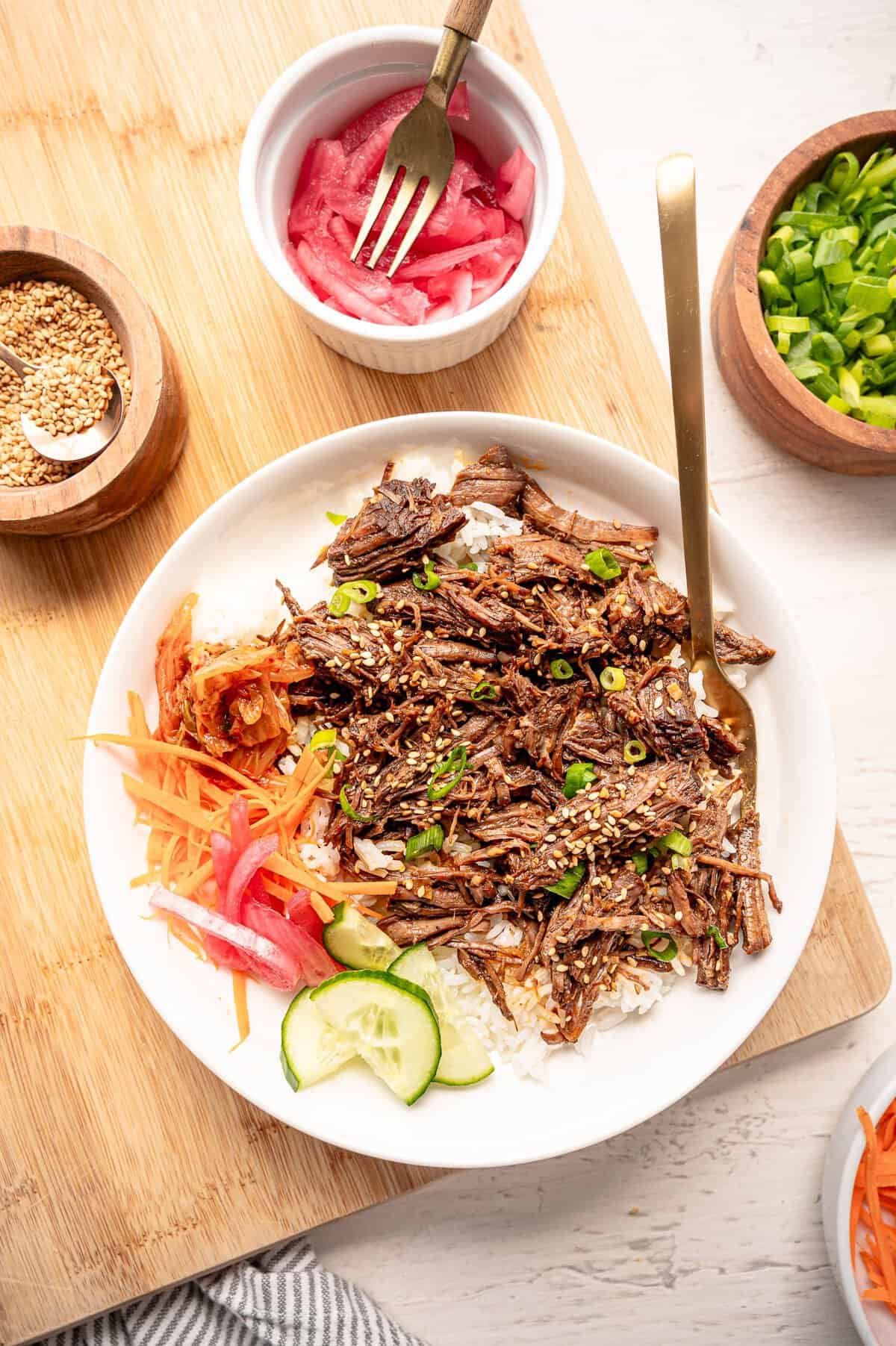 Korean beef on a plate with white rice, cucumber slices, shredded carrots, pickled red onions, and Kimchi on the side.