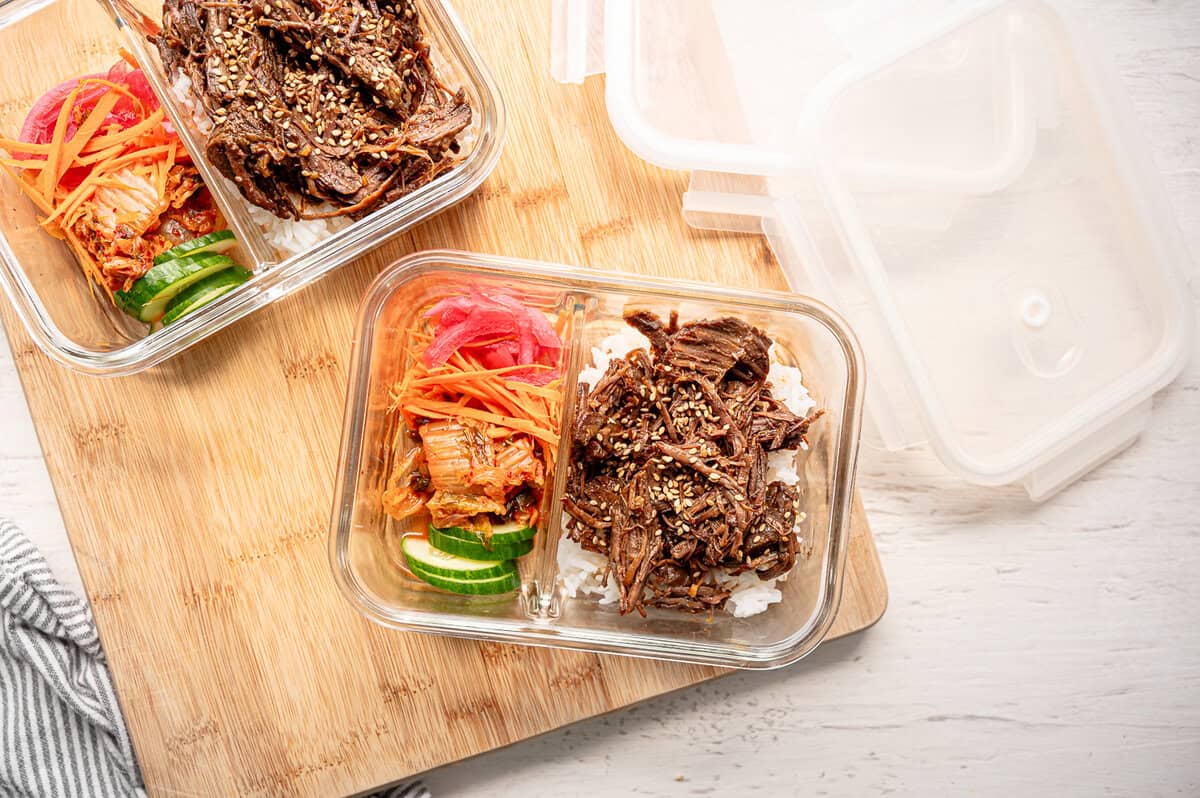 A divided glass meal prep container with shredded Korean beef and rice on one side and various fresh veggies on the other ready to take for lunch or dinner.