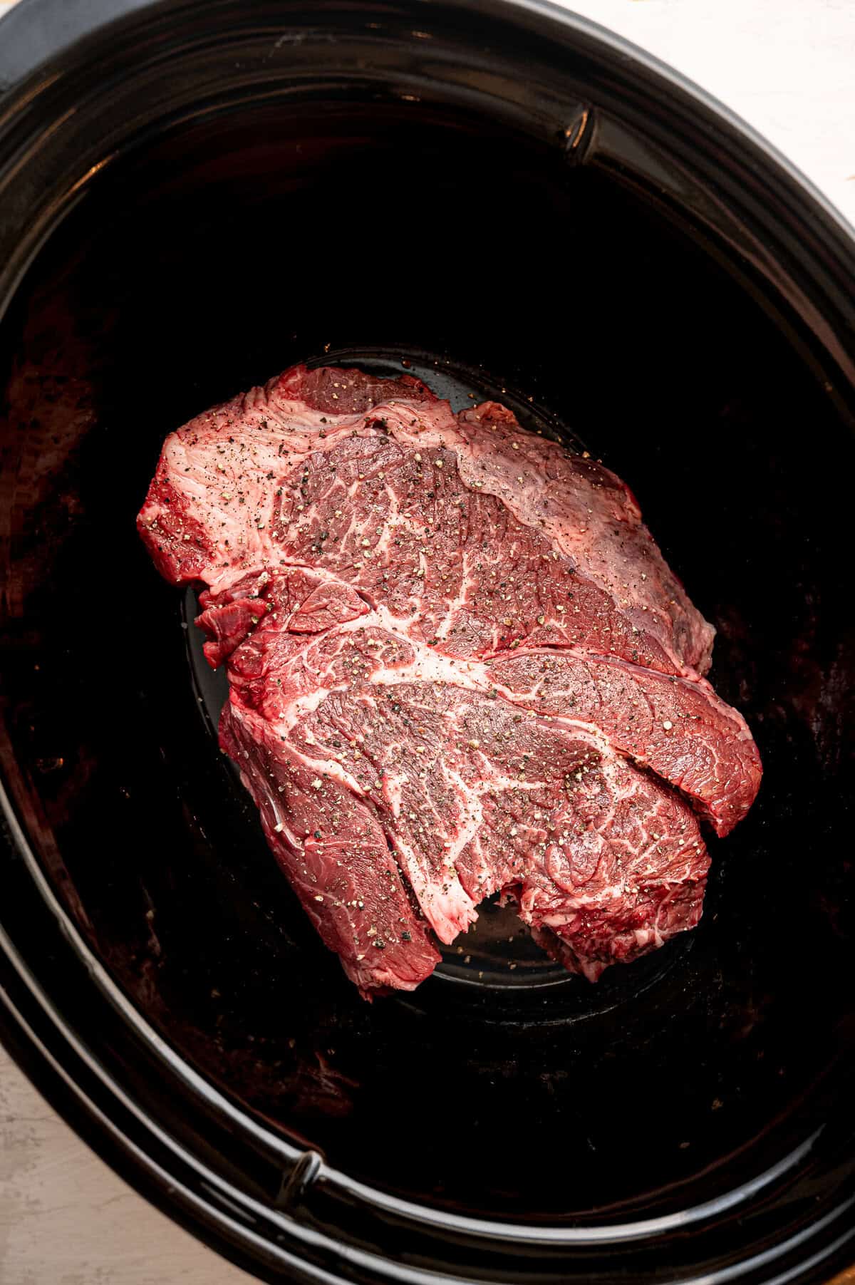 A raw roast in a slow cooker with pepper on it.