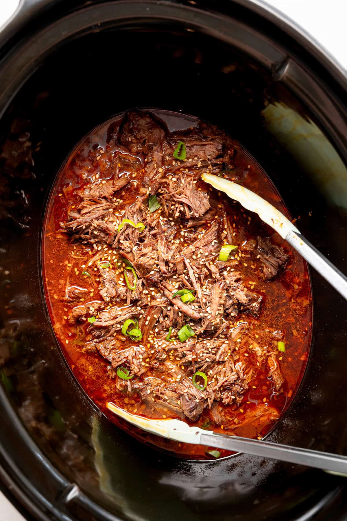 Tongs being used to pick up a portion of Korean beef from a slow cooker liner.